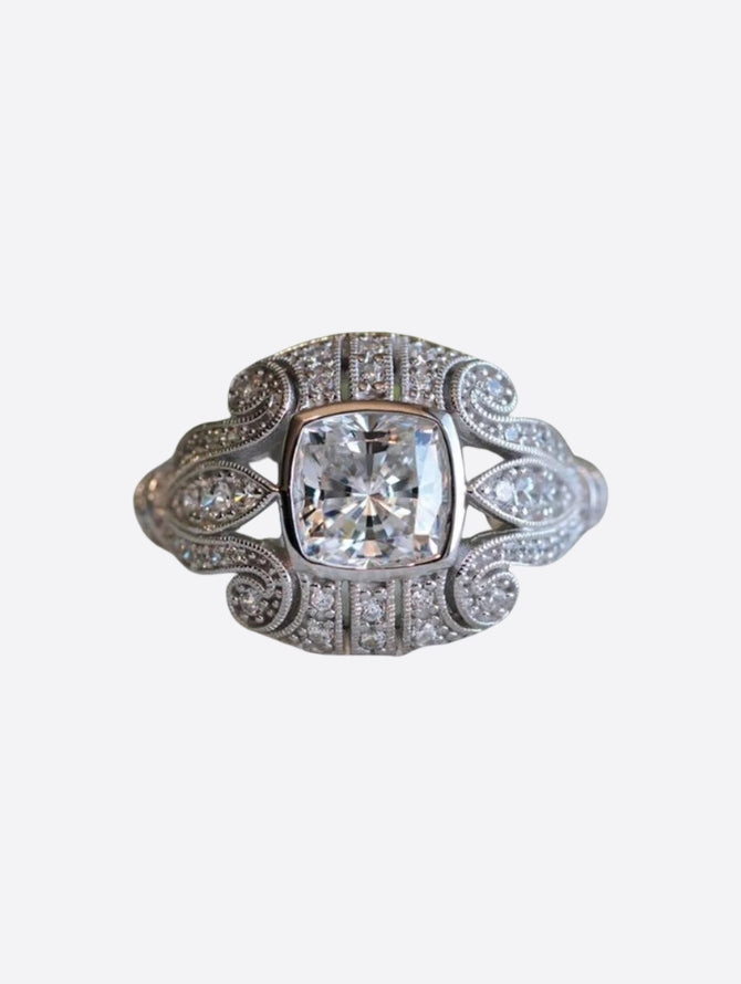A vintage-inspired silver ring featuring a large square-cut clear gemstone in the centre, surrounded by intricate filigree detailing and smaller clear gemstones, creating an elegant and ornate design.