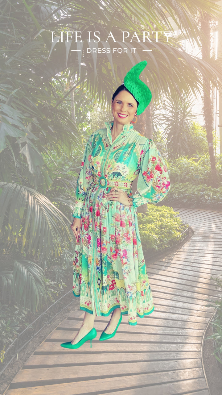 A woman stands on a wooden walkway surrounded by lush greenery, wearing a vibrant green dress with intricate floral patterns, matching green high heels, and a unique green hat. The dress has a high collar and a belted waist. 