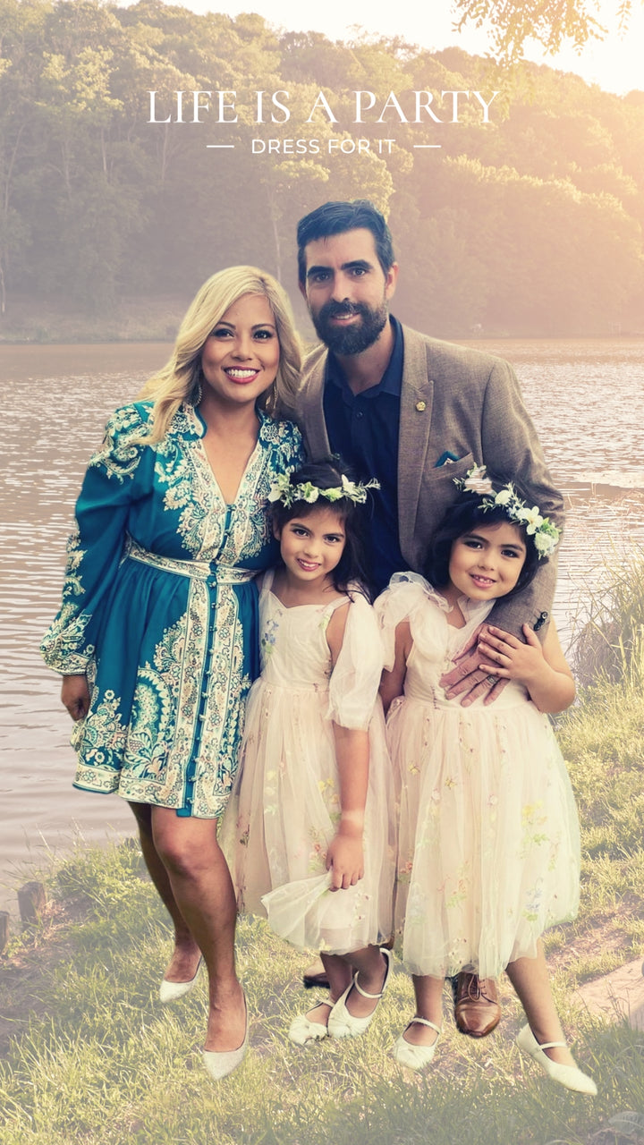 A woman stands beside a man and two young girls by a lakeside, all smiling. The woman is wearing a teal dress with white intricate patterns, accessorised with a matching belt. The man is dressed in a beige blazer with a dark shirt. The two young girls are wearing matching white dresses with floral crowns. The background features a serene lake and lush green trees. 