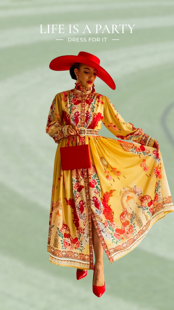 A woman poses in a flowing yellow dress adorned with red floral designs, accessorized with a large red hat, red heels, and a red handbag. The dress features a high collar and detailed embroidery. 